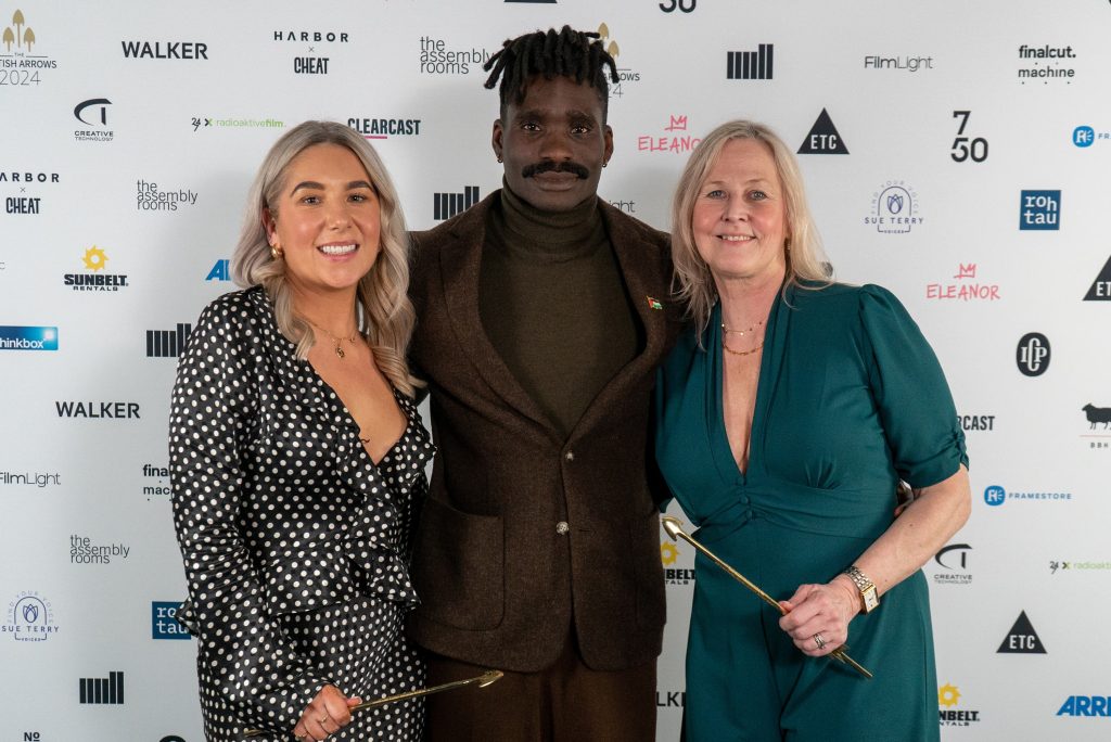 3 people stand and smile in front of a white board covered in various logos A woman in a spotty dress, a man in a brown suit and a woman in a teal dress, who holds a gold arrow award.